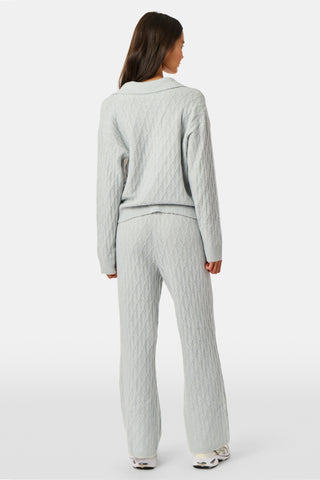 A woman wearing a light gray Cable Knots Rocio Sweater by Roller Rabbit, featuring a textured knit and V-neck, paired with matching knit pants, stands against a plain white background.
