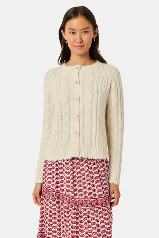 The Ruby Cardigan by REVOLUTION KNITS is a cream-colored, alpaca blend cable-knit cardigan with a crew neck and button front, worn over a red and white patterned skirt.