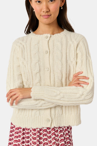The Ruby Cardigan by REVOLUTION KNITS is a cream-colored, alpaca blend cable-knit cardigan with a crew neck and button front, worn over a red and white patterned skirt.