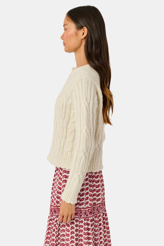 The Ruby Cardigan by REVOLUTION KNITS is a cream-colored, alpaca blend cable-knit cardigan with a crew neck and button front, worn over a red and white patterned skirt.