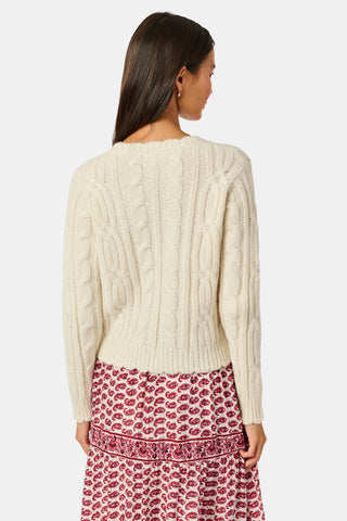 The Ruby Cardigan by REVOLUTION KNITS is a cream-colored, alpaca blend cable-knit cardigan with a crew neck and button front, worn over a red and white patterned skirt.