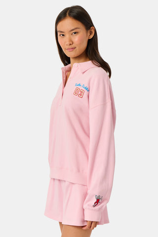 A young woman in the Roller Rabbit Game Day Rugby Sweatshirt and cap smiles while holding a football on a grassy field with trees and buildings in the background.