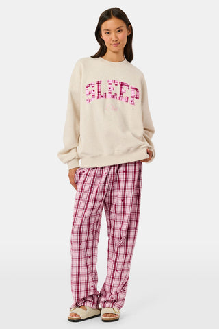 A woman wears the ARILCONFEX Sleep Club Andie Sweatshirt, an oatmeal crewneck with SLEEP in pink plaid appliqué across the chest and Club embroidered below it in pink script.