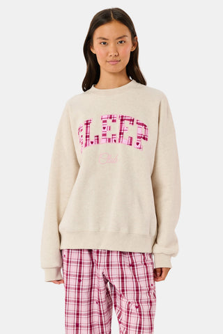 A woman wears the ARILCONFEX Sleep Club Andie Sweatshirt, an oatmeal crewneck with SLEEP in pink plaid appliqué across the chest and Club embroidered below it in pink script.