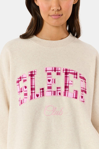 A woman wears the ARILCONFEX Sleep Club Andie Sweatshirt, an oatmeal crewneck with SLEEP in pink plaid appliqué across the chest and Club embroidered below it in pink script.