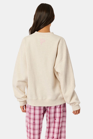 A woman wears the ARILCONFEX Sleep Club Andie Sweatshirt, an oatmeal crewneck with SLEEP in pink plaid appliqué across the chest and Club embroidered below it in pink script.