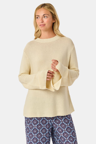 A woman wearing the Roller Rabbit Soleil Sweater, a cream cashmere blend pullover with wide sleeves, paired with patterned blue and white pants, stands against a plain white background.