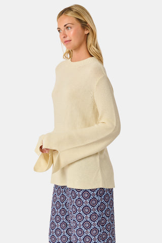 A woman wearing the Roller Rabbit Soleil Sweater, a cream cashmere blend pullover with wide sleeves, paired with patterned blue and white pants, stands against a plain white background.