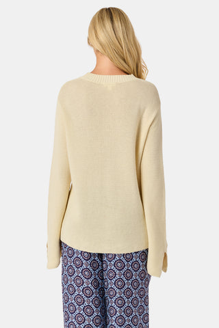 A woman wearing the Roller Rabbit Soleil Sweater, a cream cashmere blend pullover with wide sleeves, paired with patterned blue and white pants, stands against a plain white background.