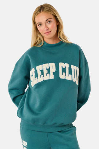 A woman wears an ARILCONFEX Sleep Club Taylor Mockneck in green with a long pale pink scarf and white shorts featuring a small red pattern.