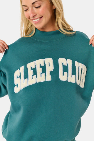 A woman wears an ARILCONFEX Sleep Club Taylor Mockneck in green with a long pale pink scarf and white shorts featuring a small red pattern.