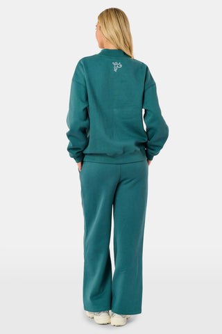 A woman wears an ARILCONFEX Sleep Club Taylor Mockneck in green with a long pale pink scarf and white shorts featuring a small red pattern.
