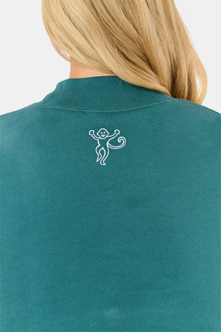 A woman wears an ARILCONFEX Sleep Club Taylor Mockneck in green with a long pale pink scarf and white shorts featuring a small red pattern.