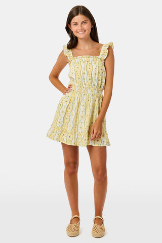 A girl wears the Roller Rabbit Kids Lillian Floral Stripe Tristan Dress, a cotton sundress with green and white stripes, floral patterns, ruffled straps, and a ruffled hem, standing with a smile and one arm relaxed at her side.