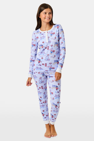 A young child with curly hair stands barefoot against a plain white background, wearing Roller Rabbit’s Kids London Monkey Pajamas—light blue long-sleeve pajamas adorned with playful, colorful patterns featuring guards, clocks, and iconic British landmarks.