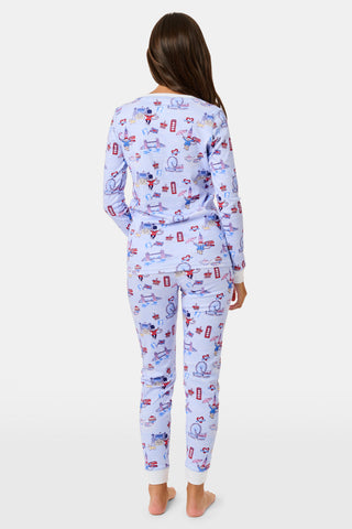 A young child with curly hair stands barefoot against a plain white background, wearing Roller Rabbit’s Kids London Monkey Pajamas—light blue long-sleeve pajamas adorned with playful, colorful patterns featuring guards, clocks, and iconic British landmarks.