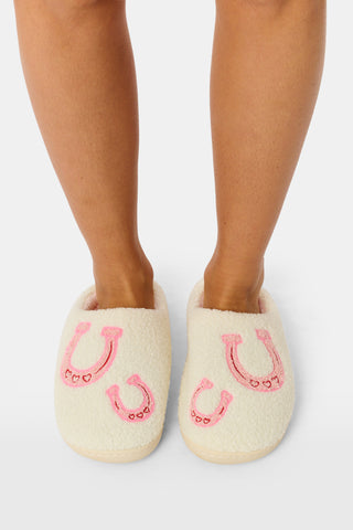 A person stands on a white background wearing Roller Rabbit Lasso Bows Fuzzy Slippers, which feature a cream-colored fuzzy finish, pink horseshoe designs, and durable rubber soles.