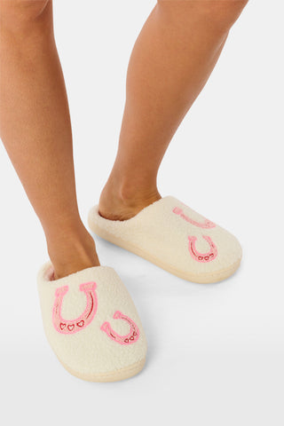A person stands on a white background wearing Roller Rabbit Lasso Bows Fuzzy Slippers, which feature a cream-colored fuzzy finish, pink horseshoe designs, and durable rubber soles.