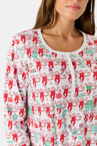A woman stands outside with her arms crossed, wearing the Roller Rabbit Alpha Chi Omega Monkey Pajamas—a white Henley top and elastic waist pajama set featuring playful red and pink monkey patterns. She holds a matching quilted bag and carries a small heart-shaped blue and white polka dot purse.