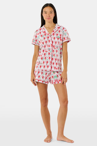 Wearing Roller Rabbit’s Alpha Chi Omega Monkey Polo Pajamas in a playful print, a woman leans against a brick wall, smiling softly as she holds two pink and white patterned notebooks and an iced coffee with a straw.