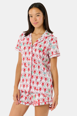 Wearing Roller Rabbit’s Alpha Chi Omega Monkey Polo Pajamas in a playful print, a woman leans against a brick wall, smiling softly as she holds two pink and white patterned notebooks and an iced coffee with a straw.