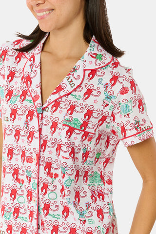 Wearing Roller Rabbit’s Alpha Chi Omega Monkey Polo Pajamas in a playful print, a woman leans against a brick wall, smiling softly as she holds two pink and white patterned notebooks and an iced coffee with a straw.