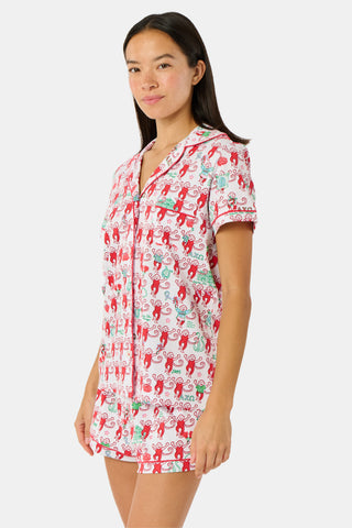 Wearing Roller Rabbit’s Alpha Chi Omega Monkey Polo Pajamas in a playful print, a woman leans against a brick wall, smiling softly as she holds two pink and white patterned notebooks and an iced coffee with a straw.