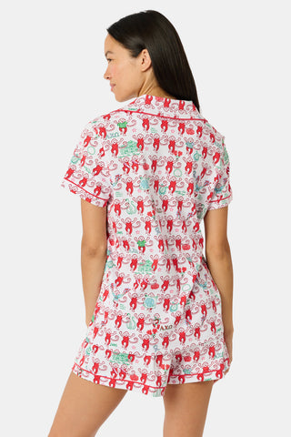 Wearing Roller Rabbit’s Alpha Chi Omega Monkey Polo Pajamas in a playful print, a woman leans against a brick wall, smiling softly as she holds two pink and white patterned notebooks and an iced coffee with a straw.