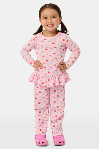 A young girl stands smiling, wearing the Roller Rabbit Kids Apple Bloom Elle Set—a pink girls’ loungewear outfit featuring an apple bloom print and a ruffled hem. She has long dark hair and is wearing light pink shoes, set against a plain white background.