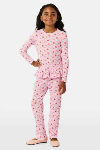 A young girl stands smiling, wearing the Roller Rabbit Kids Apple Bloom Elle Set—a pink girls’ loungewear outfit featuring an apple bloom print and a ruffled hem. She has long dark hair and is wearing light pink shoes, set against a plain white background.
