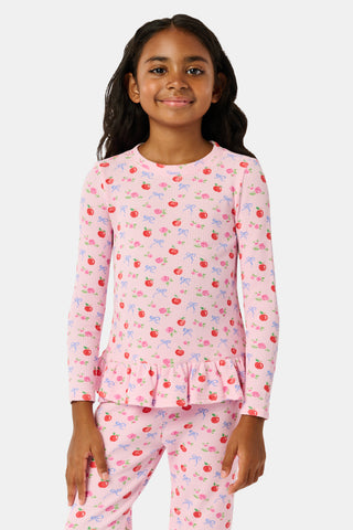 A young girl stands smiling, wearing the Roller Rabbit Kids Apple Bloom Elle Set—a pink girls’ loungewear outfit featuring an apple bloom print and a ruffled hem. She has long dark hair and is wearing light pink shoes, set against a plain white background.