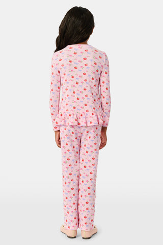 A young girl stands smiling, wearing the Roller Rabbit Kids Apple Bloom Elle Set—a pink girls’ loungewear outfit featuring an apple bloom print and a ruffled hem. She has long dark hair and is wearing light pink shoes, set against a plain white background.