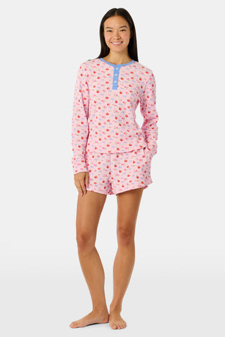 A woman wears the Roller Rabbit Apple Bloom Rosemary Set—a soft Pima cotton pajama ensemble featuring a pink long-sleeve top with a blue button placket and matching shorts, both adorned with a delicate red and pink apple bloom print. She stands with one hand in her pocket, gazing to the side.