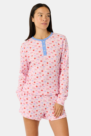 A young girl stands smiling, wearing the Roller Rabbit Kids Apple Bloom Elle Set—a pink girls’ loungewear outfit featuring an apple bloom print and a ruffled hem. She has long dark hair and is wearing light pink shoes, set against a plain white background.