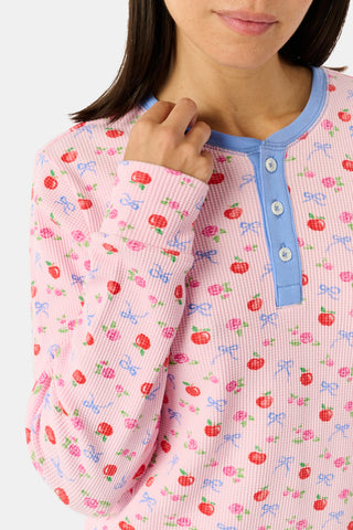 A woman wears the Roller Rabbit Apple Bloom Rosemary Set—a soft Pima cotton pajama ensemble featuring a pink long-sleeve top with a blue button placket and matching shorts, both adorned with a delicate red and pink apple bloom print. She stands with one hand in her pocket, gazing to the side.