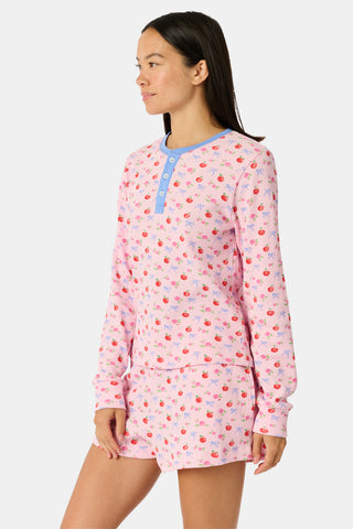 A woman wears the Roller Rabbit Apple Bloom Rosemary Set—a soft Pima cotton pajama ensemble featuring a pink long-sleeve top with a blue button placket and matching shorts, both adorned with a delicate red and pink apple bloom print. She stands with one hand in her pocket, gazing to the side.