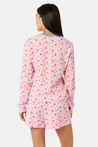 A woman wears the Roller Rabbit Apple Bloom Rosemary Set—a soft Pima cotton pajama ensemble featuring a pink long-sleeve top with a blue button placket and matching shorts, both adorned with a delicate red and pink apple bloom print. She stands with one hand in her pocket, gazing to the side.