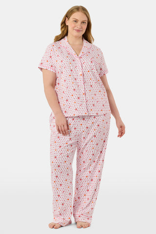 A woman in the Roller Rabbit Autumn Hearts Fiora Pajamas and brown boots sits on a wooden fence, smiling and resting one arm on a tree branch in a sunny outdoor setting, evoking the coziness of fall sleepwear.