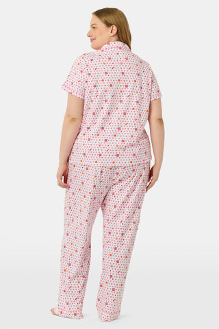 A woman in the Roller Rabbit Autumn Hearts Fiora Pajamas and brown boots sits on a wooden fence, smiling and resting one arm on a tree branch in a sunny outdoor setting, evoking the coziness of fall sleepwear.