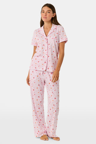 A woman in the Roller Rabbit Autumn Hearts Fiora Pajamas and brown boots sits on a wooden fence, smiling and resting one arm on a tree branch in a sunny outdoor setting, evoking the coziness of fall sleepwear.