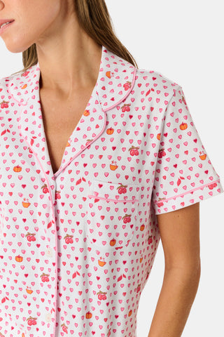 A woman in the Roller Rabbit Autumn Hearts Fiora Pajamas and brown boots sits on a wooden fence, smiling and resting one arm on a tree branch in a sunny outdoor setting, evoking the coziness of fall sleepwear.