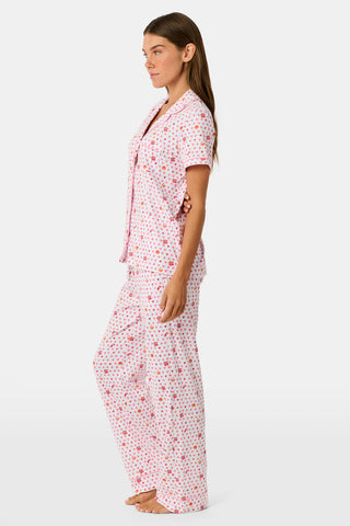 A woman in the Roller Rabbit Autumn Hearts Fiora Pajamas and brown boots sits on a wooden fence, smiling and resting one arm on a tree branch in a sunny outdoor setting, evoking the coziness of fall sleepwear.