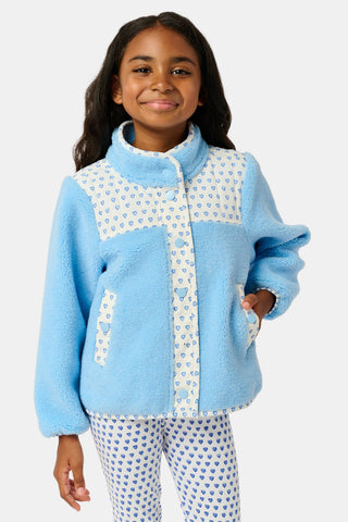A young girl stands and smiles against a plain background, wearing the Roller Rabbit Kids Hearts Lizzie Fleece Jacket in light blue with white patterned details, paired with matching white leggings adorned with blue hearts. This cozy outerwear features soft fleece for comfort and warmth.