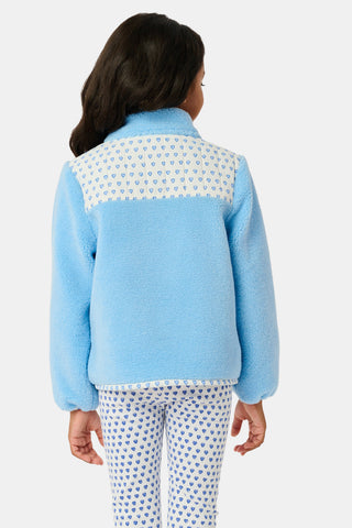 A young girl stands and smiles against a plain background, wearing the Roller Rabbit Kids Hearts Lizzie Fleece Jacket in light blue with white patterned details, paired with matching white leggings adorned with blue hearts. This cozy outerwear features soft fleece for comfort and warmth.