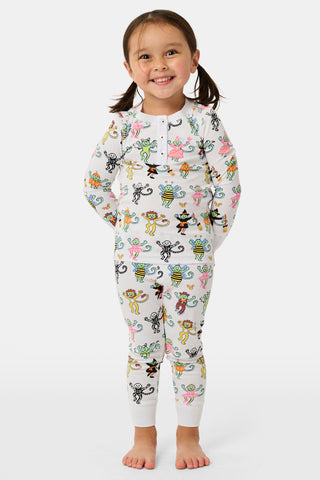 A smiling child in Roller Rabbit’s Kids Boo Crew Pajamas, crafted from soft Pima cotton, sits with arms raised on outdoor steps among women in coordinating patterned pajamas, all sharing a cheerful and relaxed moment together.