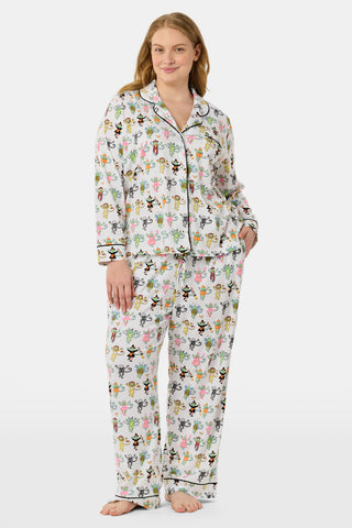 A woman stands barefoot against a plain white background, wearing the Roller Rabbit Boo Crew Long Sleeve Polo Pajamas, which feature a colorful cartoon bee and flower pattern on white Pima cotton with long sleeves and button-up styling.
