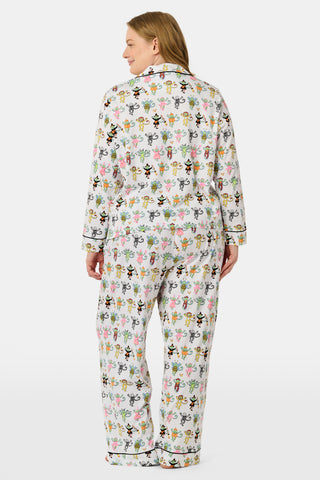 A woman stands barefoot against a plain white background, wearing the Roller Rabbit Boo Crew Long Sleeve Polo Pajamas, which feature a colorful cartoon bee and flower pattern on white Pima cotton with long sleeves and button-up styling.
