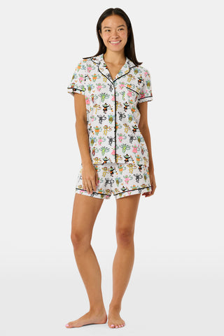 A smiling child in Roller Rabbit’s Kids Boo Crew Pajamas, crafted from soft Pima cotton, sits with arms raised on outdoor steps among women in coordinating patterned pajamas, all sharing a cheerful and relaxed moment together.