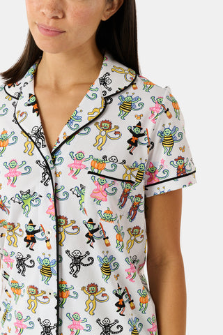 A smiling child in Roller Rabbit’s Kids Boo Crew Pajamas, crafted from soft Pima cotton, sits with arms raised on outdoor steps among women in coordinating patterned pajamas, all sharing a cheerful and relaxed moment together.
