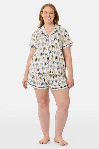 A smiling child in Roller Rabbit’s Kids Boo Crew Pajamas, crafted from soft Pima cotton, sits with arms raised on outdoor steps among women in coordinating patterned pajamas, all sharing a cheerful and relaxed moment together.